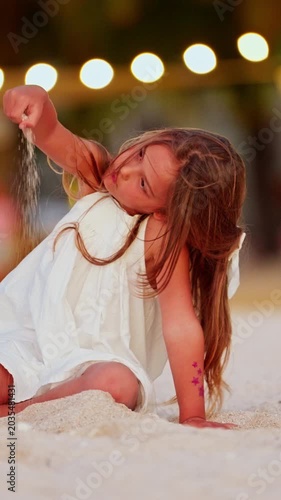 A little beautiful girl in a white sundress has fun on the beach spilling sand through fingers of a tropical island on vacation at sunset