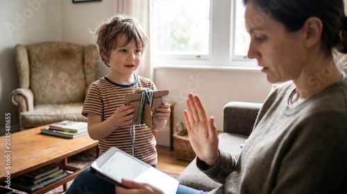 A young child holding a craft looks at an adult woman making a stop gesture. The woman is engrossed in a tablet, highlighting parental distraction.