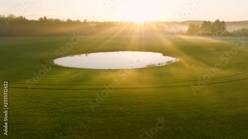 Aerial slow motion tracking pan of circular pond reflecting sunrise over green field in countryside