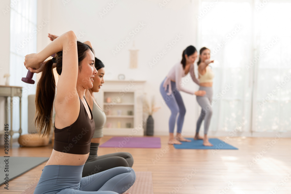 Fototapeta premium Women exercise in a bright studio with weights and yoga mats during a fitness class focusing on strength and flexibility on a sunny day