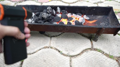 First person view of hand using portable blower to ignite charcoal in home brazier. Outdoor cooking scene with glowing coals, fire, smoke, and heat.