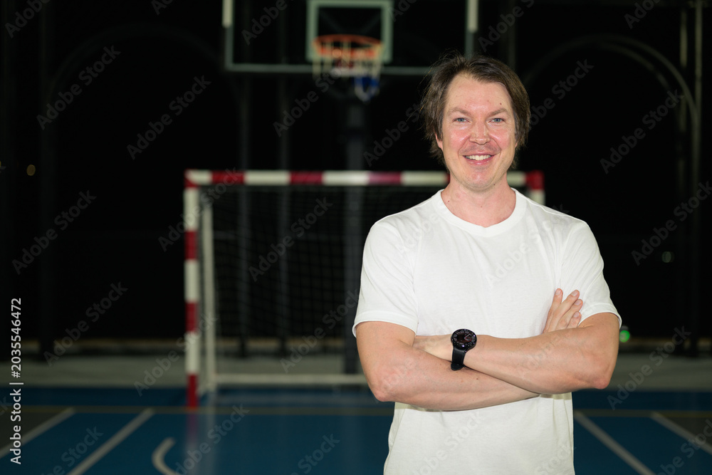Naklejka premium Smiling athletic Caucasian man in his late 30s posing with arms crossed on horizontal outdoor sports court at night