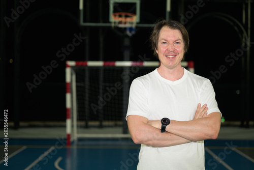 Smiling athletic Caucasian man in his late 30s posing with arms crossed on horizontal outdoor sports court at night
