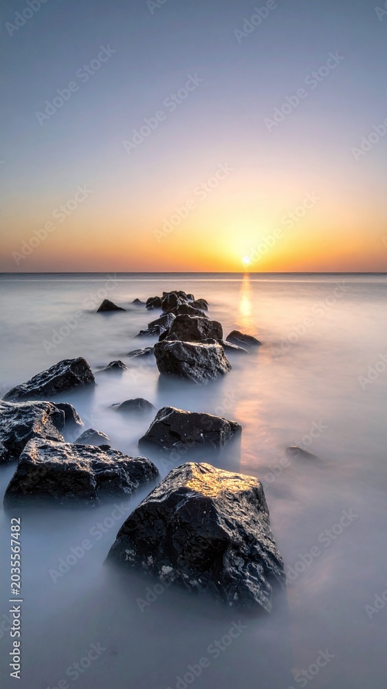 Obraz premium Serene long exposure captures dark rocks leading into a golden sunrise over a misty ocean. This peaceful landscape is perfect for wellness, meditation and travel marketing content.