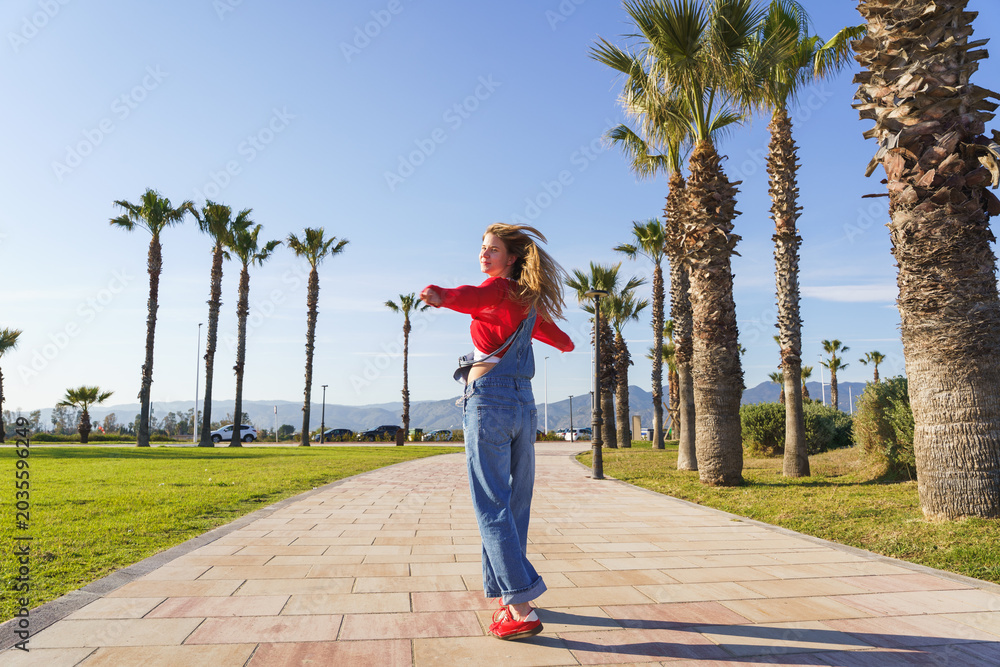 Fototapeta premium Young woman rotating happily on a path lined with palm trees under clear sky