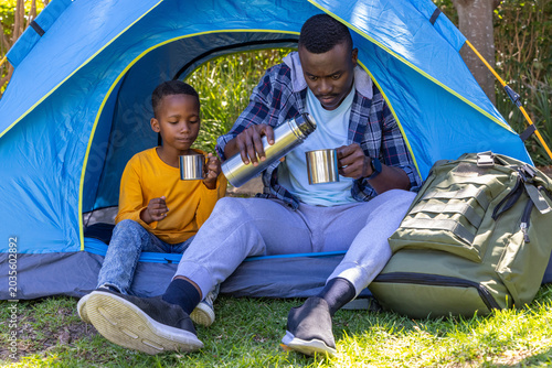 African American father and son sitting at blue dome tent entrance pouring thermos into metal cups