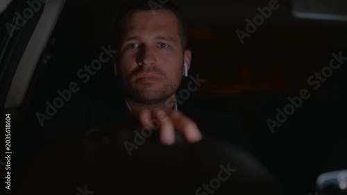 Young man driving car at night while using wireless headphones, listening the music. Right hand drive car. Traveling, trip, night life concept. Real time