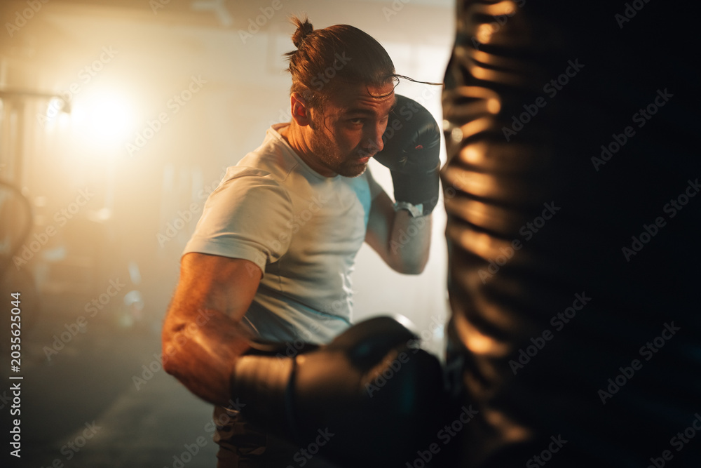 Fototapeta premium Focused man exercising with boxing gloves on a heavy bag in a dimly lit fitness studio, improving health and stamina