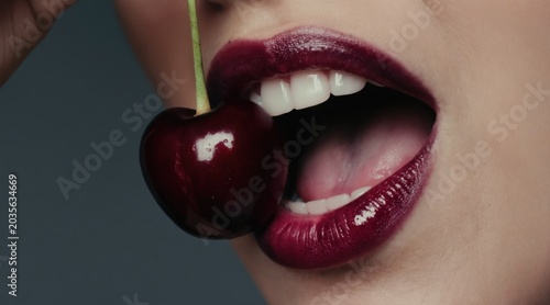 Woman open mouth with glossy deep red lipstick and white teeth, holding dark red cherry by stem. She bites cherry with sensuous expression, food close-up.