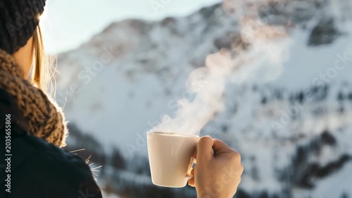 A person holds a steaming cup of coffee in front of a snowy mountain view. Sunlight shines on the snow, and steam rises from the mug. The scene captures a cold morning with warmth from the drink