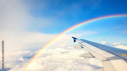 The camera shows a clear view from the airplane wing above fluffy clouds. A rainbow appears in the blue sky, creating a colorful cloud landscape during flight