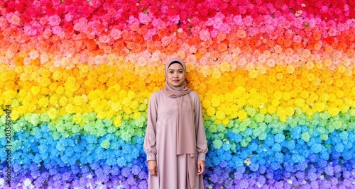 Woman wearing hijab standing before vibrant ombre floral wall, multicolored flowers, spring celebration, traditional fashion, faith and nature
