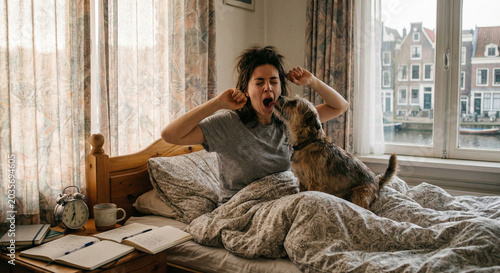Tired woman yawning and stretching in bed while pet dog licks her face during sunny morning awakening.