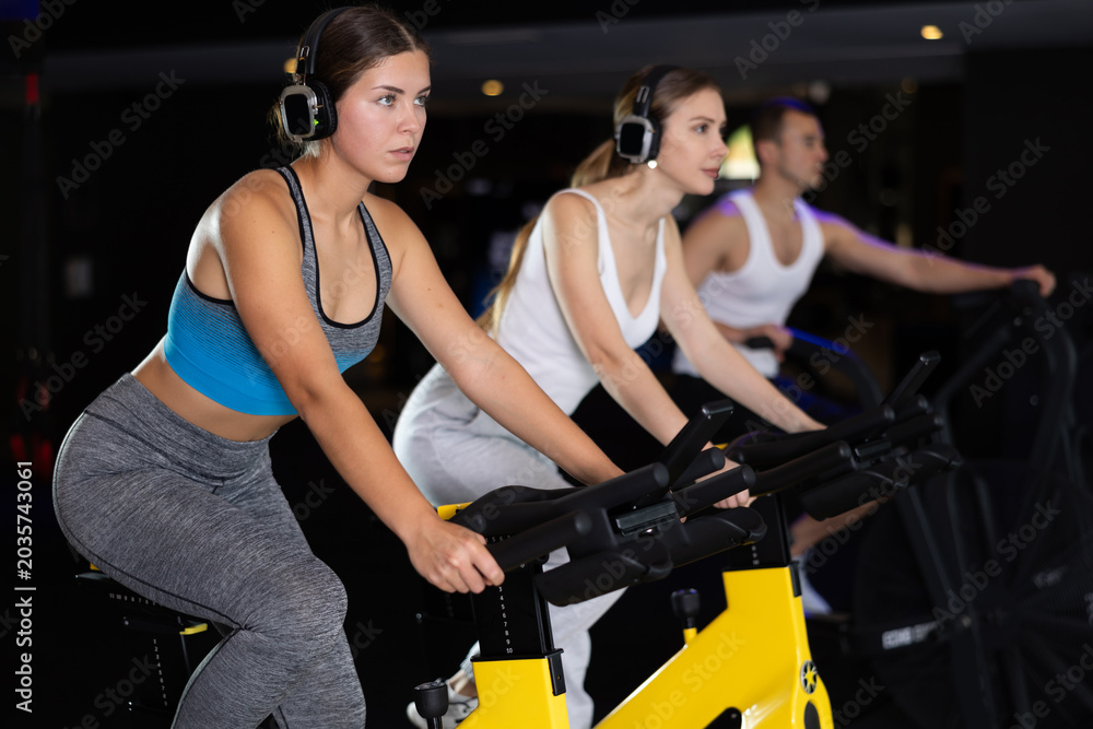Fototapeta premium Girl in earphone spend time in gym, improve physical shape with exercises. Female sport studio visitor listen to music and uses exercise bike to pump leg muscles. Strength and motivation, fitness goal