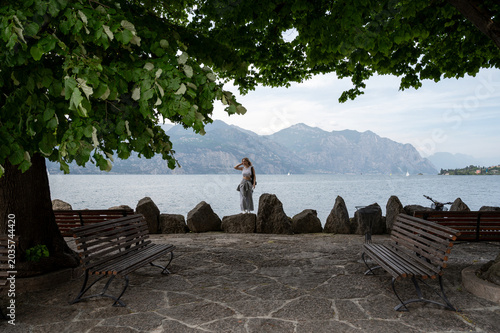 Woman standing by a lakeside viewpoint on Lake Garda, Italy, with benches, trees, mountains, and calm water in the background.