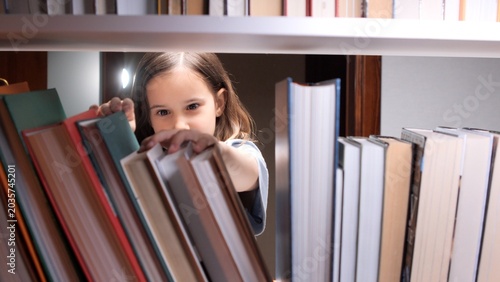 Photography Young girl with long brown hair reaching for a book on a crowded library shelf,