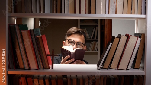Obraz na płótnie Man in eyeglasses reading a holy book intently between bookshelves with a wooden
