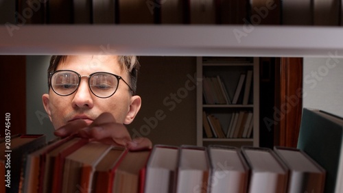 Slika na platnu Man wearing glasses looking carefully at books on a library shelf, reaching for