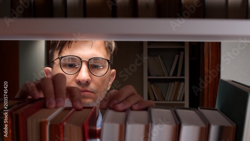 Cuadro en lienzo Man in eyeglasses reaching for a book on a library shelf, focused on selecting r