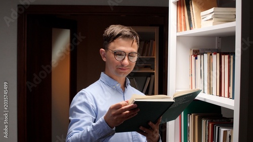 Slika na platnu Man wearing glasses and a light blue shirt standing next to a bookshelf, holding