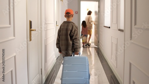 Family checking out of hotel, mother and daughter walk ahead while smiling son pulls his suitcase down bright hallway after a relaxing vacation stay, ready for the next destination