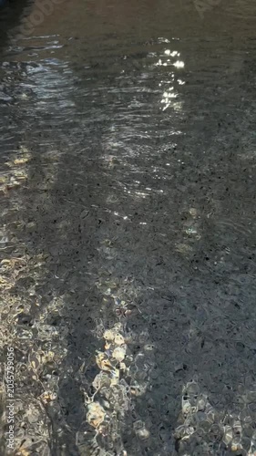 Slow motion static handheld pan of sunlight reflections shimmering across shallow pebbled shoreline water at daytime beach