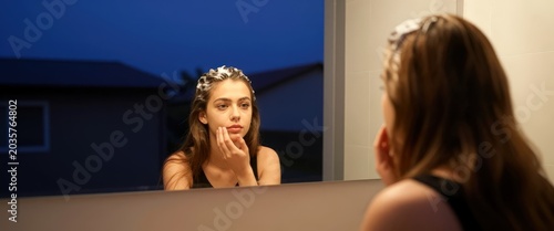 Young woman with foam on head looking in bathroom mirror at twilight. Use: personal care, beauty routine, self-care.