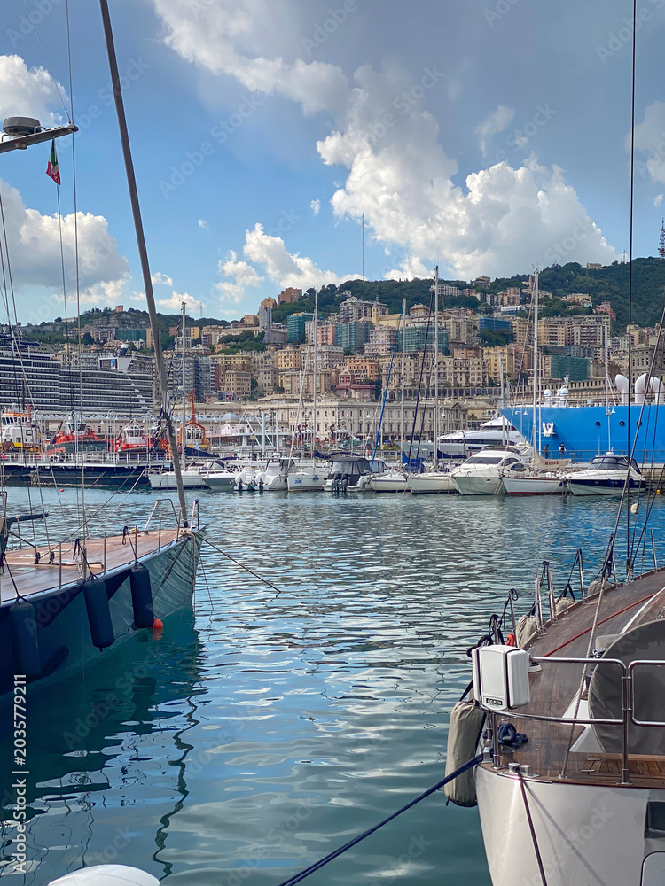 Obraz premium Yachts and sailboats moored in Porto Antico marina with a cruise ship and Genoa skyline against blue sky with clouds, Italy