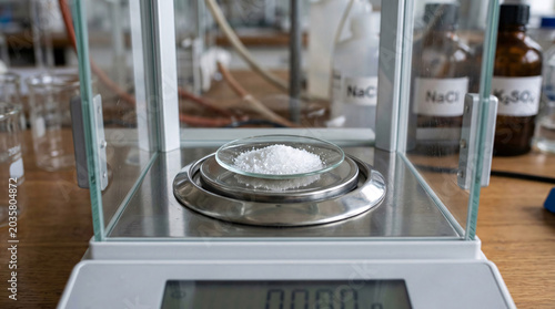 Laboratory digital scale with sodium chloride sample and chemical reagent bottles in background.
