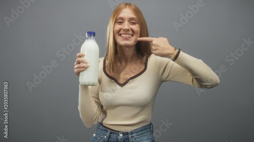 Woman redhead holding milk bottle with left hand and pointing finger to cheek while smiling in studio setting under soft light; wholesome joy.