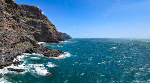 Photography Rocky coastline with dark volcanic rocks and clear blue ocean waves lapping agai
