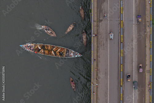 Dhaka, Bangladesh - 25 May 2026: Aerial view of the Buriganga River with a large boat transporting cattle alongside smaller wooden vessels near a busy bridge with rickshaws.