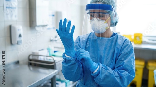 A healthcare worker is donning blue protective gear in a clinical setting. The worker is wearing blue scrubs, nitrile gloves, a face shield, and a surgical mask.