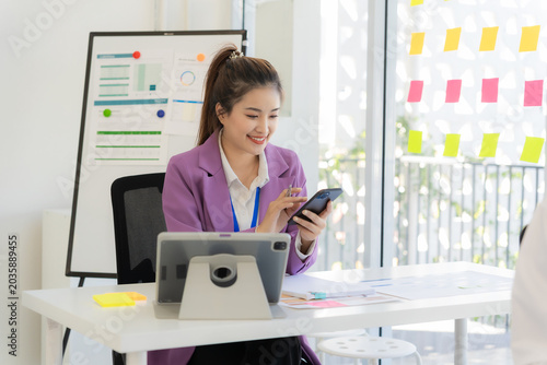 A happy Asian businesswoman chats on her smartphone while working on a digital tablet at her desk, holding a cup of coffee and calling a client in the workspace.