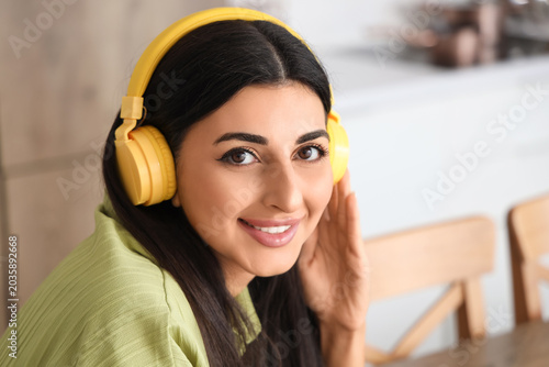 Quadro su tela Beautiful young woman with modern headphones listening to music in kitchen