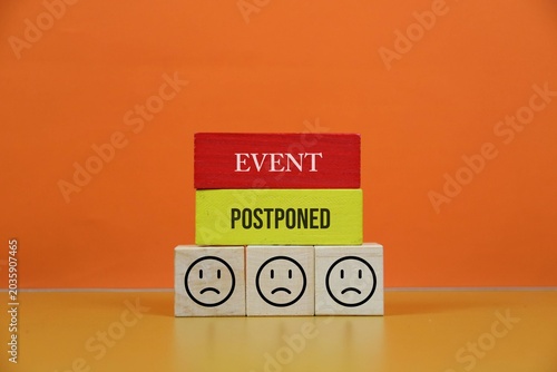 Postponed Event Announcement With Wooden Blocks and Sad Faces Representing Cancellation and Disappointment, with an orange background.
