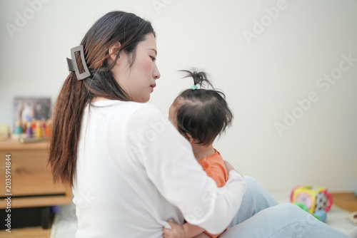 Mother holds child close, offering comfort. Soft light highlights tender moment between them. Child rests head on mother's shoulder, trusting. Background holds toys and framed photos