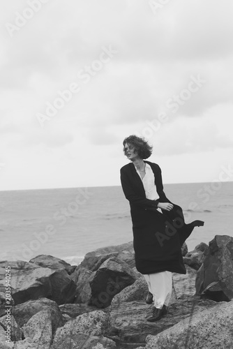 Fotografi Woman in a dark coat stands on rugged rocks by the sea, wind tousling her hair,