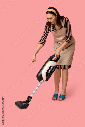 Portrait of worried young housewife in apron with vacuum cleaner pointing at ...