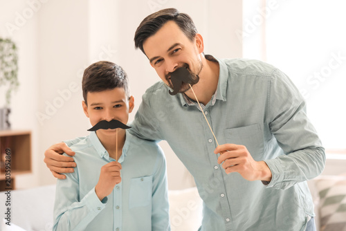 Portrait of father and his little son with paper mustache at home