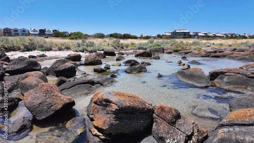 Clear shallow tidal pools shimmer among dark rust-coloured boulders at Jawbone Marine Sanctuary in Williamstown, Melbourne, Australia, with suburban residential buildings visible along the horizon