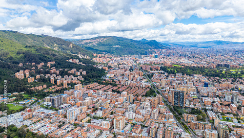 Vast cityscape of Bogotá with residential buildings and green mountains in the background under a cloudy sky.