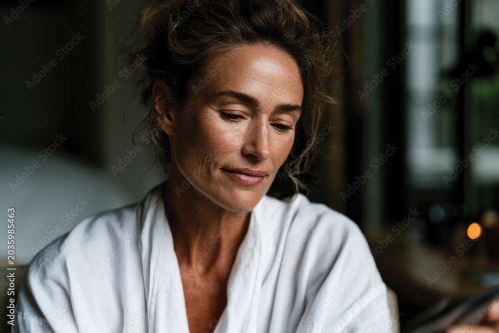 Fototapeta premium A serene moment captured of a woman in a soft robe, highlighting her calm demeanor as she engages with her phone in a cozy indoor setting, surrounded by soft lighting.