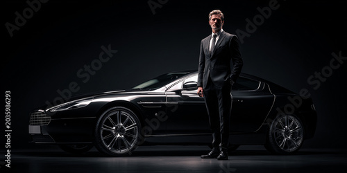Stylish man in suit stands confidently next to sleek black luxury car in dark setting, exuding elegance and sophistication
