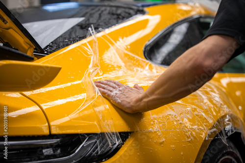 Inside a luxury car workshop, a technician smooths wet film on a yellow sports car rear fender, taillight and raised rear wing visible, under bright overhead lights.