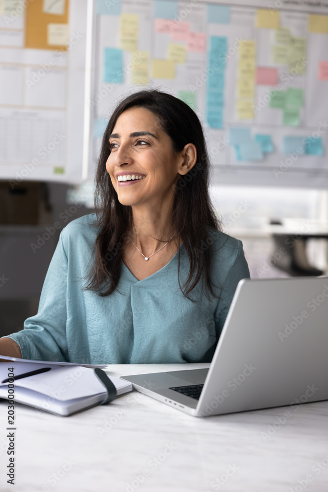 Obraz premium Happy professional woman sits at desk with open laptop holding documents, review data, feel satisfied with project progress, sales growth, take break, thinks on perspectives, career and opportunities
