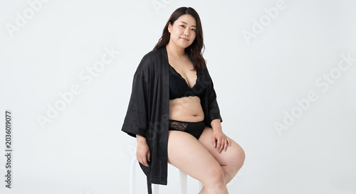 A woman sits on a stool in front of a white background, wearing a black robe and black lingerie. She is looking at the camera with a slight smile.