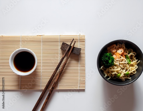 Asian noodle bowl with vegetables soy sauce chopsticks on bamboo mat top view on white background minimalist composition