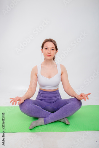 Full length portrait of young athletic woman sitting in lotus position on yoga mat isolated on white background.
