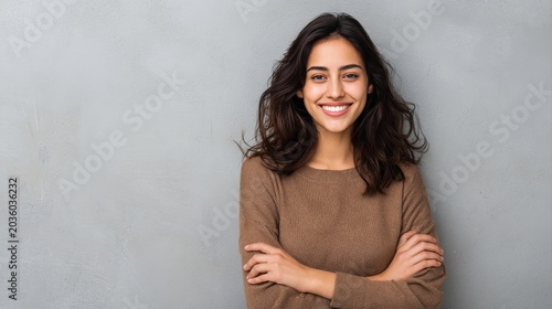 Portrait of a young latin woman with pleasant smile and crossed arms isolated on grey wall with copy space. Beautiful girl with folded arms looking at camera against grey wall. Cheerful hispanic woman